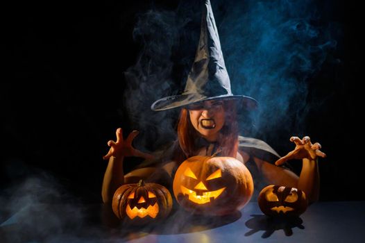 The evil witch casts a spell on pumpkins. Portrait of a woman in a carnival halloween costume in the dark.