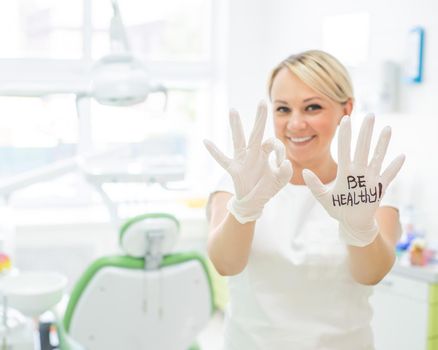 Friendly female dentist smiling and showing okay sign with fingers. Blonde doctor in gloves in the office in the medical center. The inscription on the hand be healthy.