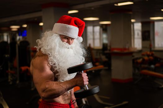 Santa claus in the gym. Muscular man with a naked torso doing exercises with dumbbells