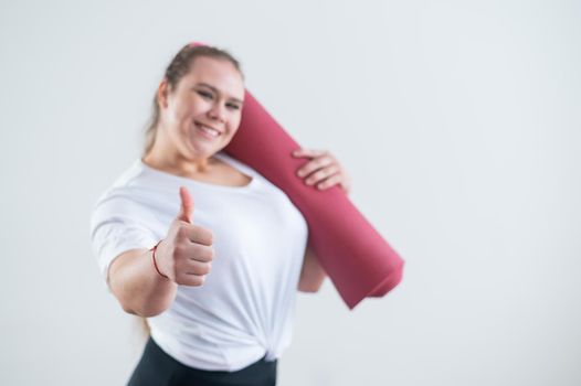 Young fat caucasian woman holding a sports mat and showing a thumb up. Charming plus size model in sportswear stands on a white background.