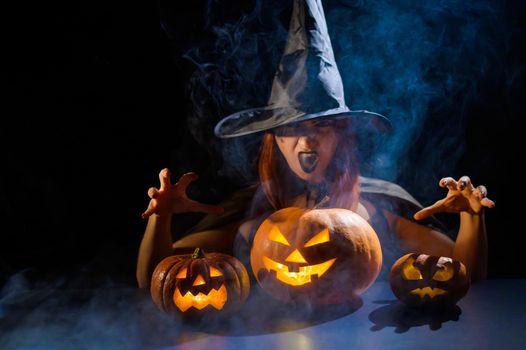 The evil witch casts a spell on pumpkins. Portrait of a woman in a carnival halloween costume in the dark.