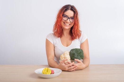 Caucasian woman prefers healthy food. Redhead girl chooses between broccoli and donuts on white background