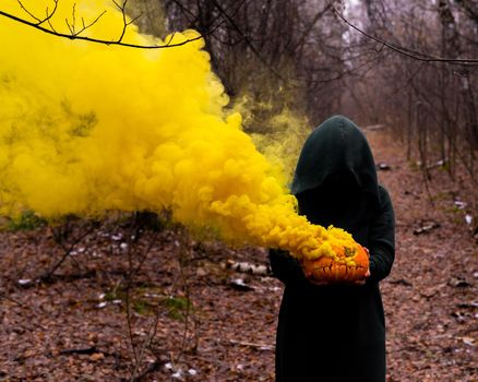 A creepy witch holds a steaming pumpkin in a deep forest. Jack o lantern emits yellow smoke for halloween.