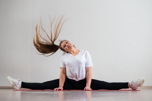 A young flexible fat woman sits in a transverse twine and waves her hair against a white background.