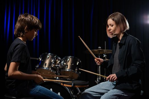 Young caucasian woman teaches a boy to play the drums in the studio on a black background. Music school student.