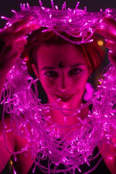 Woman with witch makeup and an earring in her nose. The girl holds a garland of flickering pink lights near her face. Light bulbs illuminate the witch's face in the dark