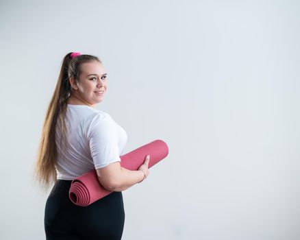 Young fat caucasian woman holding a sport mat. Charming plus size model in sportswear stands on a white background.