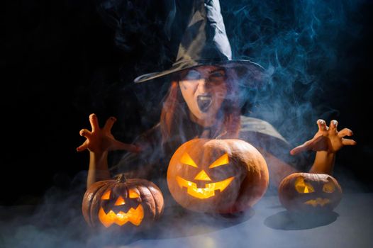 An ominous witch in a hat conjures over a jack-o-lantern. Traditional halloween characters. Mystical fog creeps over pumpkins with carved terrible faces. An evil witch casts a spell