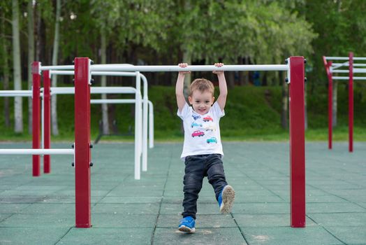 A little boy learns to pull up on a horizontal bar in the open air. The child is hanging on the uneven bars