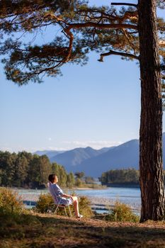 A woman is sitting on a folding chair on the bank of a mountain river on a nice, warm day under a large tree. A calm and quiet place to relax and reflect. Equipment and a tourist's rest.