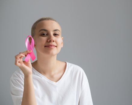 Young woman with short hair wearing a white t-shirt holding a pink ribbon as a symbol of breast cancer on a white background