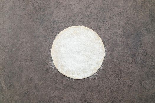 Thin homemade pizza dough with scattered wheat flour on dark concrete background.