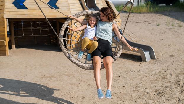 Mom and daughter swing on a round swing. Caucasian woman and little girl have fun on the playground