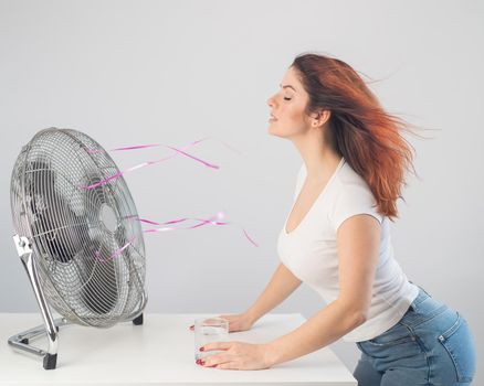 A red-haired Caucasian woman chills by the electric fan and drinks a cold drink. Climate control in the apartment.