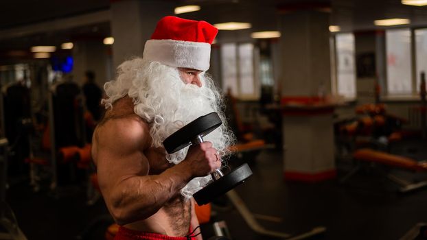 Santa claus in the gym. Muscular man with a naked torso doing exercises with dumbbells