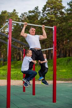 Caucasian man and two boys doing exercises outdoors. The father pulls himself up on the horizontal bar with his sons on the playground
