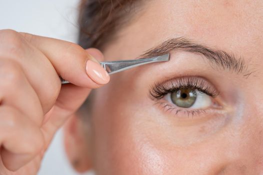 Close-up portrait of a caucasian woman doing eyebrow correction herself with tweezers.