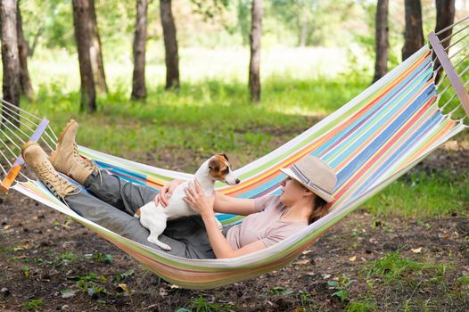 Caucasian woman lies in a hammock with Jack Russell Terrier dog in a pine forest.