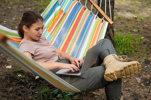 Caucasian woman working on laptop while sitting in a hammock in the forest. Girl uses a wireless computer on a hike