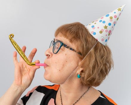 Portrait of a smiling elderly woman in a festive cap holding a whistle tongue on a white background.