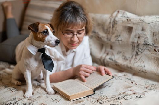 An elderly caucasian woman is lying on a sofa with a smart dog jack russell terrier wearing glasses and a tie and reading a book