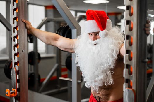 Santa claus in the gym. Muscular man with a naked torso doing exercises with dumbbells