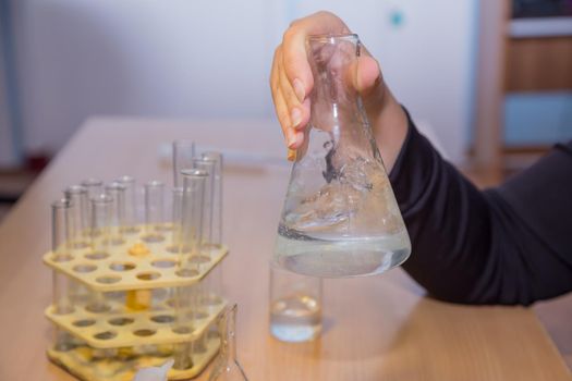 Close-up hands of a student shake up a chemical liquid in a flask. A schoolboy performs a task at the workplace. The concept of children's education, teaching knowledge, skills and abilities.