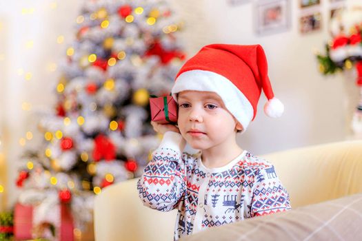 Boy at the Christmas tree. The boy folded his hands.. Cute baby . Holiday. Article about new year and Christmas. Colorful spruce. Garland. Holiday decoration and decor . Santa hat.