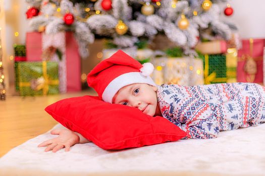 Boy at the Christmas tree. The boy folded his hands.. Cute baby . Holiday. Article about new year and Christmas. Colorful spruce. Garland. Holiday decoration and decor . Santa hat.