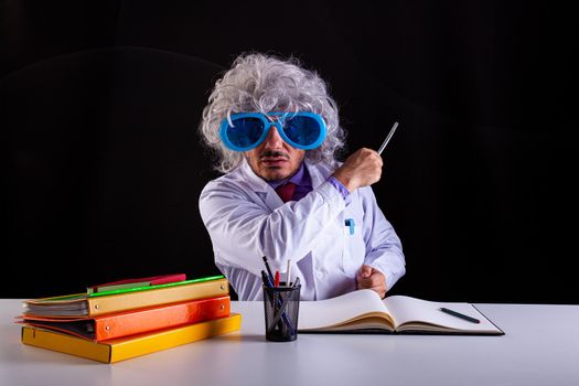 Crazy science teacher in white coat with unkempt hair in funny eye glasses sitting at the desk holding a wand to point at the blackboard