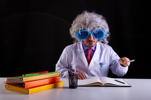 Crazy science teacher in white coat with unkempt hair in funny eye glasses sitting at the desk