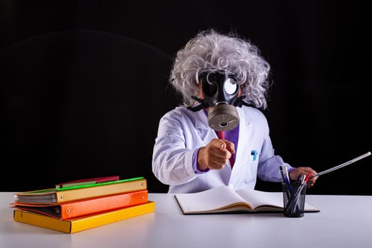 Crazy science teacher in white coat with unkempt hair sitting at the desk wears a gas mask