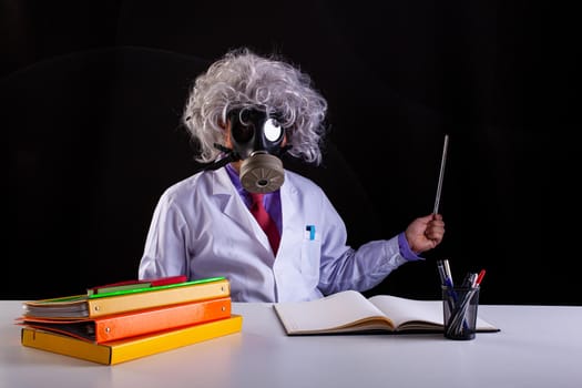 Crazy science teacher in white coat with unkempt hair sitting at the desk wears a gas mask