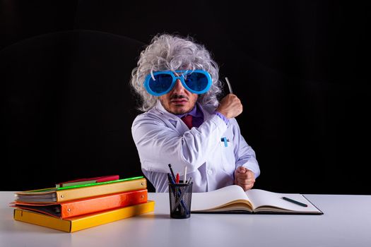 Crazy science teacher in white coat with unkempt hair in funny eye glasses sitting at the desk holding a wand to point at the blackboard