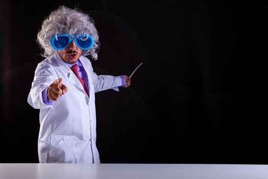 Angry science teacher in white coat with unkempt hair in funny eye glasses points with his finger on black background