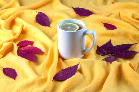 Autumn composition of red dry leaves laid out on a bright yellow crumpled cloth and a ceramic mug with hot tea with lemon. Selective focus.