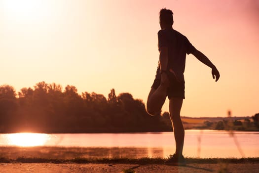 A young man trains outdoors against the backdrop of a beautiful lake at sunset, the athlete warms up, does exercises on the legs before running.