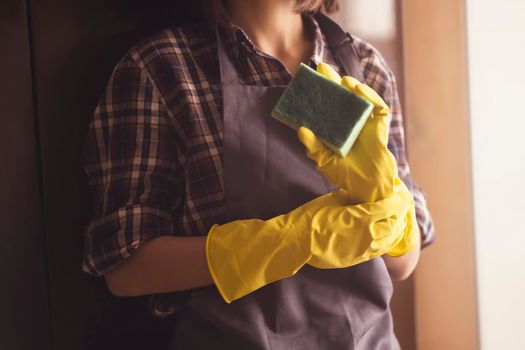Young female hands in yellow rubber gloves hold a new green sponge for cleaning house and wiping surfaces. A female housekeeper in a plaid shirt and a gray apron holds a rag and plans to disinfect.