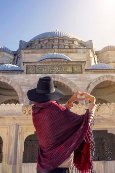 A young traveler in a hat and red poncho folds her hands in the shape of a heart against the background of an ancient blue mosque in Istanbul, Turkey. Girl tourist travels to Arab countries.