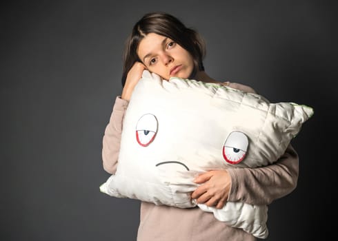 Cute sad young girl hugs a white pillow with painted eyes and face. The woman is unhappy with early waking up, sleep problems and insomnia.