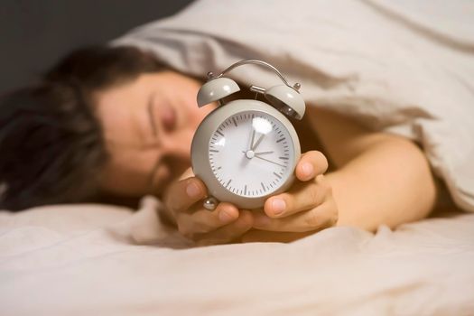 Hands of a young man from under the blankets hold a retro vintage alarm clock in gray. The person holds a clock, need to wake up.