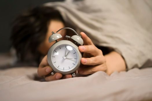 Hands of a young man from under the blankets hold a retro vintage alarm clock in gray. The person holds a clock, need to wake up.