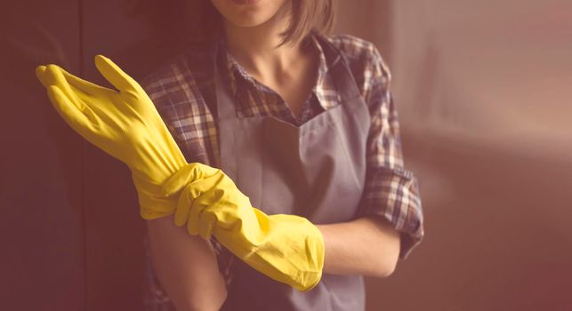 A young girl in a plaid shirt and apron puts yellow rubber gloves on her hands to start cleaning her house and create comfort. Housekeeper with gloves doing disinfection.