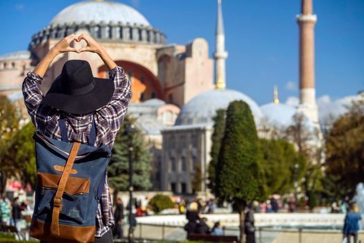 Young traveler in a hat and stylish shirt folds her hands in the shape of a heart against the background of an ancient Hagia Sophia mosque in Istanbul, Turkey. Girl tourist travels to Arab countries.