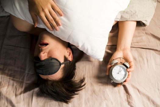 A young funny man sleeps soundly early in the morning, hugs a pillow and holds a vintage alarm clock in his hands, the sleep mask has slipped from his eyes.