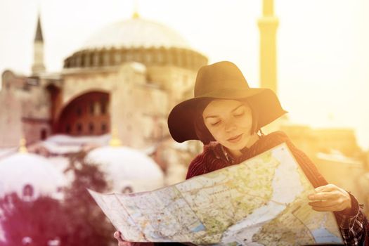 A young girl in a hat looks at a map of Istanbul on a sunny day. Beautiful ancient Hagia Sophia Mosque on the background. Traveling to Arab countries.