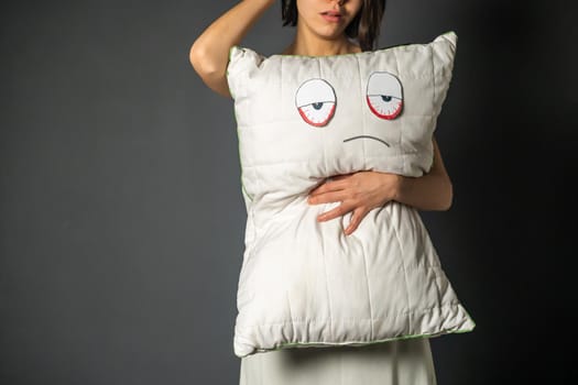 Cute sad young girl hugs a white pillow with painted eyes and face. The woman is unhappy with early waking up, sleep problems and insomnia.