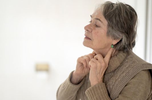 An elderly woman applies an anti-aging An elderly woman puts fingers on the lymph nodes and looks at the mirror. Takes care, monitors thyroid hormones and the health of the endocrine system.