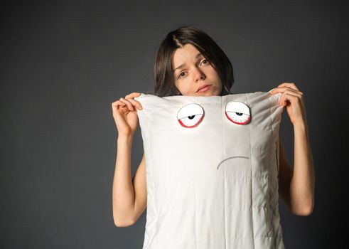 Young girl with a disgruntled face holds a pillow, on which a displeased face and tired eyes are drawn. Creative idea about sleep problems, insomnia and the importance of quality orthopedic materials.