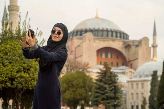 A young smiling Muslim girl in a hijab and stylish black glasses makes a selfie against the background of the Hagia Sophia mosque. Tourism in Istanbul, Turkey